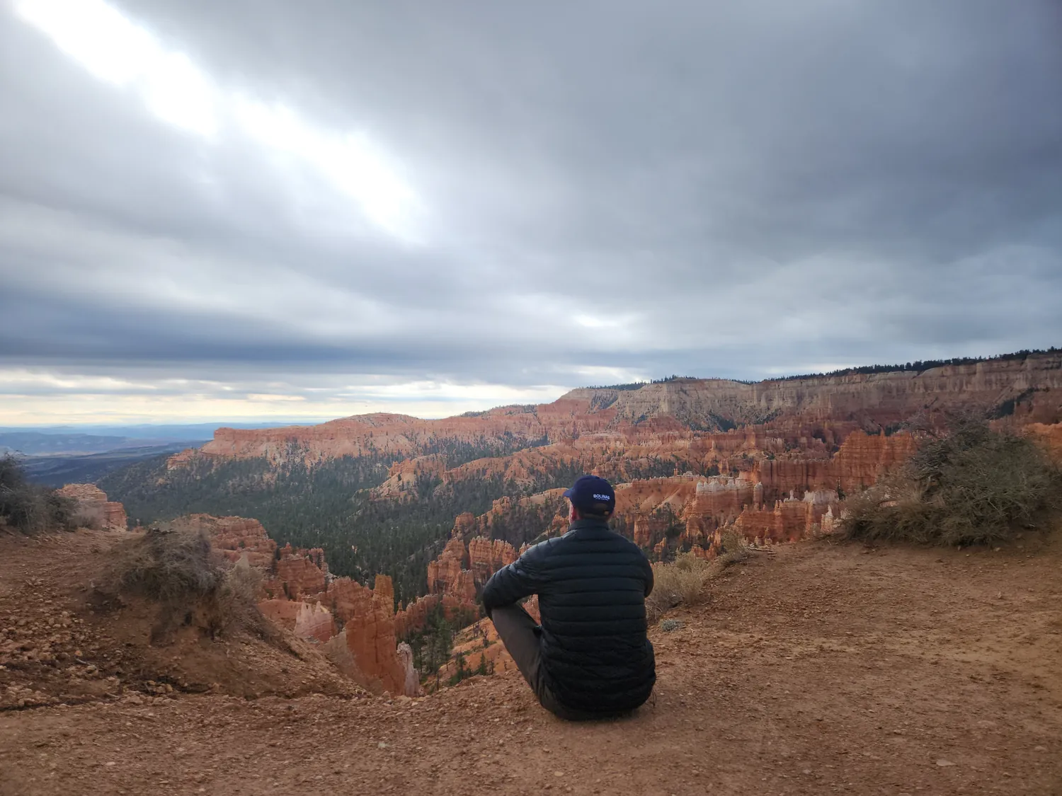 A hiker sits quietly on the edge of a canyon rim, looking out over red-orange rock spires and forest under a cloudy sky.