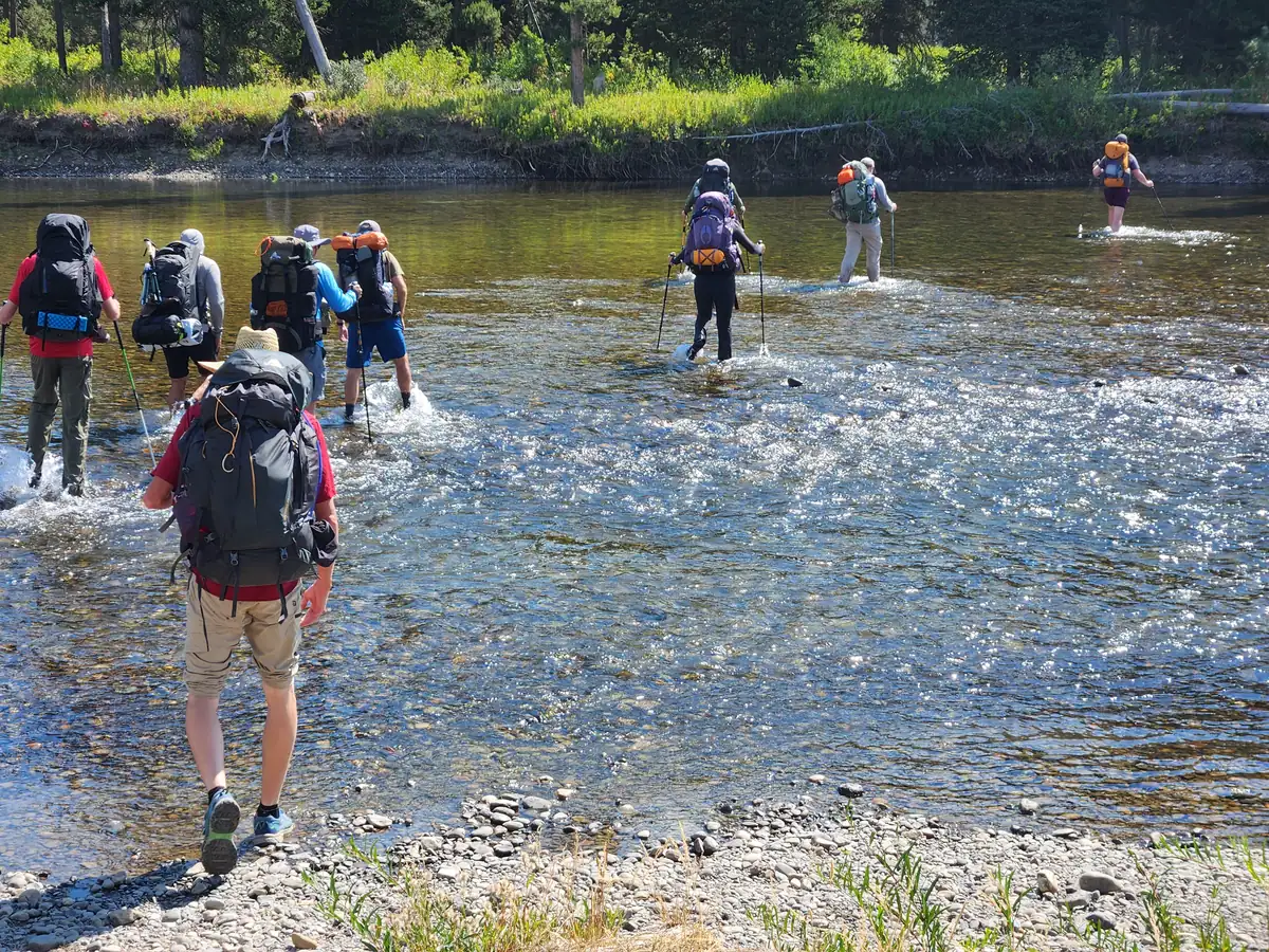 A group of backpackers wading single file across a wide, shallow river, seen from behind.